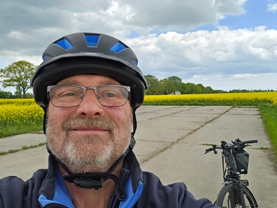 Eine Person mit Fahrradhelm macht ein Selfie auf einem betonierten Weg in ländlicher Umgebung. Hinter der Person steht ein Fahrrad. Links und rechts des Weges blühen gelbe Felder, darüber ein wolkiger Himmel.