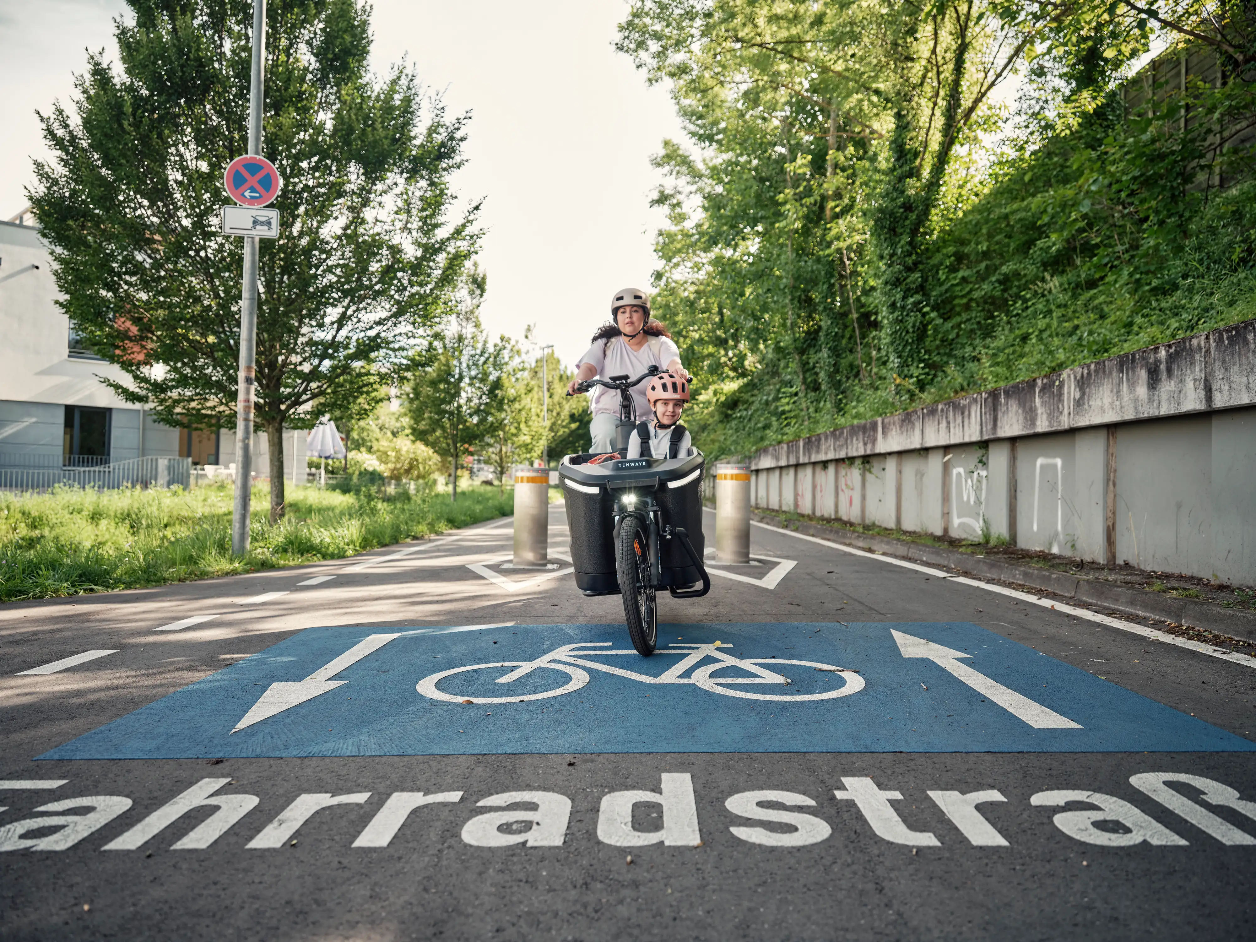 Mama mit Kind im Lastenrad fährt auf Fahrradstrasse zwischen Pollern hindurch Mama mit Kind im Lastenrad fährt auf Fahrradstrasse zwischen Pollern hindurch